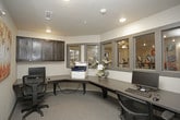 This single-floor business center is thoughtfully designed to provide a functional and comfortable workspace. The room features an L-shaped built-in desk with ample surface area, accommodating a desktop computer workstation and a full-size multifunction printer and copier. Dark-finished upper cabinetry above the desk provides additional storage, while the neutral-toned carpet and light gray walls create a clean, professional atmosphere. A colorful autumn birch tree canvas adds a decorative touch to the wall, complementing the understated interior palette. Multiple windows along one wall overlook an adjacent fitness center, allowing natural light to filter into the space while maintaining a visual connection to the surrounding amenities. A secondary workstation near the glass-paneled entry door offers an additional computer setup with direct sightlines into the fitness area. The business center is accessed through a clearly marked glass door, reinforcing the open and welcoming character of the space. A wall-mounted electrical panel is discreetly integrated into the room. Overall, the business center is a practical, well-appointed space suited for work, printing, and general productivity needs.
