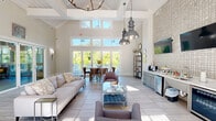 Located in Asheville, NC, this single-floor open-concept space is defined by its dramatic vaulted, beam-accented ceiling clad in white shiplap, which lends the interior a sense of generous vertical volume. Light-toned large-format tile flooring extends throughout, reflecting abundant natural light that pours in through a wall of floor-to-ceiling French doors and clerestory windows along the rear facade. These doors open directly onto an outdoor deck surrounded by lush greenery, seamlessly connecting the interior to the natural landscape.

The main living area features a comfortable seating arrangement anchored by a neutral sofa and accent chairs, centered around a glass-top coffee table. Adjacent to the lounge area, a dining setup with a wood table and upholstered chairs provides a casual gathering spot. Overhead, a combination of an antique-style chandelier and oversized industrial pendant lights add character and warmth to the bright, airy palette.

A dedicated refreshment station along one wall showcases a textured mosaic tile accent wall, wall-mounted television, built-in cabinetry with a wet bar, and a beverage refrigerator, making the space well-suited for hosting. A decorative open shelving unit and framed artwork contribute to the welcoming, curated atmosphere throughout this versatile communal retreat.