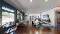 Located at 7838 Huebner Rd in San Antonio, Texas, this single-floor community clubhouse is thoughtfully designed to offer residents a variety of amenity spaces within one cohesive building. The entrance features striking floor-to-ceiling windows with teal-painted frames flanking a set of solid wood double doors, flooding the space with natural light and creating an inviting first impression. Rich, dark wood-look tile flooring runs throughout, complementing the warm architectural tones.

The leasing and reception area is highlighted by an impressive coffered ceiling with dark wood insets, pendant lighting, and symmetrically arranged workstations. A round glass-top table with a gold base serves as a centerpiece. Adjacent to the reception zone, an open lounge area offers comfortable seating, a wall-mounted display, and decorative furnishings arranged beneath arched passageways that connect the various areas seamlessly.

A dedicated dining nook features tufted teal banquette seating, patterned accent walls, and circular wall mirrors, providing an intimate social setting. The full community kitchen is equipped with stainless steel appliances, granite countertops, warm wood cabinetry, and a decorative tile backsplash. The space also includes a fitness center with exercise equipment and a game room complete with a billiards table, rounding out this versatile and well-designed community hub.