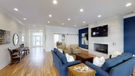 This single-floor clubhouse is thoughtfully designed to provide a comfortable and inviting gathering space for residents and guests. The main lounge area is anchored by a striking whitewashed stone fireplace paired with a wall-mounted flat-screen TV, set against a bold navy blue accent wall that adds a modern, sophisticated touch. Recessed lighting throughout the ceiling ensures the space is well-lit, complementing the warm natural light that filters in through large windows and glass entry doors visible at the far end of the room. The flooring consists of light hardwood planks that run the length of the space, contributing to a bright and airy atmosphere. Seating is arranged in a sociable configuration, featuring navy blue sofas and neutral-toned armchairs centered around a rustic wooden coffee table, creating a cozy yet stylish setting. A sliding barn door adds a charming architectural detail while providing flexible room separation. Beyond the lounge, a dining area is visible, suggesting additional space for communal meals or gatherings. Clean white trim, crown molding, and carefully chosen furnishings complete the polished, community-focused aesthetic of this well-appointed clubhouse.