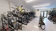 This 620-square-foot bike room in Marlborough, MA, is a purpose-built facility designed to accommodate a large number of bicycles. The space features a clean, well-lit interior with bright fluorescent lighting and white walls, creating a safe and visible environment. The room is equipped with multiple bike storage solutions, including vertical and horizontal racks, maximizing the use of available space.

The facility boasts a concrete floor, ideal for durability and easy maintenance. Various types of bicycles are accommodated, from mountain bikes to road bikes and even children's bicycles, catering to diverse cycling needs. The storage systems appear to include both floor-mounted and wall-mounted options, allowing for flexible arrangement and efficient use of vertical space.

Access to the room is via a glass door, visible in some images, which likely provides security while allowing natural light to enter. The room also features modern amenities such as a ceiling-mounted projector or security camera, enhancing the space's functionality or security.

A fire extinguisher box is visible, indicating attention to safety measures. The overall layout of the room appears well-organized, with clear pathways between bike racks for easy access and navigation. This dedicated bike room offers a secure, convenient solution for bicycle storage, likely serving residents or employees of a larger complex or building.
