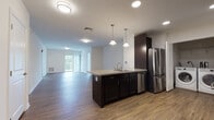 This well-appointed single-floor residence at 702 Pleasant Valley Rd N in Groton, Connecticut offers a thoughtfully designed open-concept layout. The kitchen serves as the centerpiece of the living space, showcasing dark espresso cabinetry, granite-look countertops, and a full suite of stainless steel appliances including a range, microwave, dishwasher, and refrigerator. A peninsula island with an undermount sink and elegant pendant lighting provides both additional prep space and a natural divide between the kitchen and the expansive living area. Durable hardwood-style flooring flows throughout the main living spaces, adding warmth and continuity. The generously sized living area is bathed in natural light from large windows and provides access to a private balcony. A long hallway connects the main living area to the private quarters, which include carpeted bedrooms featuring large windows with peaceful wooded views. The bathroom is finished with classic black-and-white hexagon tile flooring, dark cabinetry, a white countertop vanity, and a walk-in shower. Clean white walls, recessed lighting, and a neutral palette throughout create a bright, modern ambiance well-suited for comfortable everyday living.