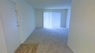 This single-floor space offers a clean and straightforward layout designed for comfortable everyday living. The main living area is open and generously proportioned, featuring soft neutral carpeting that extends throughout and light-toned walls that contribute to a bright, airy atmosphere. A large window dressed with vertical blinds anchors the primary living space, allowing abundant natural light to filter in and illuminate the room. The transition between the carpeted living area and an adjacent section with warm wood-tone flooring adds visual interest and helps delineate functional zones within the space. The walls are finished in a smooth, understated neutral palette, keeping the interior feeling fresh and adaptable to a variety of furnishing styles. The overall ambiance is calm and uncluttered, offering a blank canvas well-suited for personalization. The straightforward single-level layout ensures easy navigation throughout, making the space practical and accessible. With its combination of natural light, neutral finishes, and versatile flooring, this space provides a functional and inviting environment ready for use.