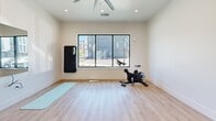 Located within a multi-use property in Sacramento, CA, this dedicated yoga and exercise room offers a clean, functional space designed to support a variety of fitness and wellness activities. The room features light wood-look flooring throughout, which adds warmth while providing a practical surface for yoga, stretching, and cardio workouts. Crisp white walls and recessed ceiling lighting create a bright, open atmosphere, while a large black-framed window along one wall invites ample natural light and views of the surrounding exterior. A ceiling fan ensures comfortable air circulation during active sessions. The space is equipped with a Matrix spin bike, a yoga mat, and a wall-mounted Reflect interactive fitness mirror, allowing for guided workouts directly within the room. A rollable exercise mat is also stored along the wall for added versatility. Striking black-framed glass doors connect this yoga room to an adjacent, larger fitness facility, providing convenient access to additional workout equipment while maintaining a visually distinct and tranquil environment. The overall ambiance is minimal and serene, thoughtfully designed to support focused movement and mindful exercise in a well-appointed community fitness setting.