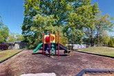 This outdoor community space offers a thoughtfully arranged set of amenities spread across a generously sized grounds. At the heart of the recreational area is a vibrant, multi-colored playground structure set within a wood chip-cushioned enclosure, bordered by low edging for safety. The playground is shaded by large, mature trees that provide a natural canopy overhead.

Adjacent to the playground, a fenced swimming pool area is visible alongside a well-kept clubhouse building. The clubhouse features a pitched roofline, classic gabled detailing, covered entryway with columns, and neatly trimmed shrubbery along its façade, giving it an inviting, residential character.

The grounds are enclosed by black iron fencing throughout, providing security while maintaining visual openness. Landscaping across the property includes a mix of mulched areas, grass lawns, ornamental shrubs, and mature deciduous trees that add seasonal color and natural shade.

A brick-accented walkway connects key areas of the property, while classic lamp posts contribute to a well-lit, community-oriented atmosphere. The two-story residential building visible in the background features brick construction, balconies, and individual unit entries, completing the cohesive character of this community setting.