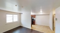 This single-floor space is designed with an open layout that seamlessly connects the living area to the kitchen. The living area features dark hardwood flooring that adds warmth and contrast to the bright, white walls and smooth white ceiling. A window along one wall allows natural light to filter into the space, keeping the interior feeling airy and well-lit. Recessed ventilation and ceiling-mounted light fixtures are visible throughout, ensuring functional comfort.

The transition from the living area to the kitchen is marked by a shift in flooring from dark hardwood to light-toned tile, clearly delineating each functional zone. The kitchen is equipped with rich, warm-toned wood cabinetry that provides ample storage, complemented by dark countertops and black appliances, including a refrigerator and range. A dedicated ceiling light fixture illuminates the kitchen area effectively.

White baseboards run throughout the space, providing a clean, finished look. The overall aesthetic is practical and straightforward, with a neutral color palette that offers versatility for personalization. The layout is efficient, making good use of the available space across the single floor.