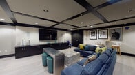 Located at 1099 NE 79th Street in Miami, this single-floor space presents a thoughtfully designed contemporary lounge environment with a cohesive, upscale aesthetic. The interior is defined by a striking coffered ceiling with bold black beams set against a neutral cream backdrop, complemented by recessed lighting that creates a warm, inviting ambiance throughout.

The main lounge area serves as the focal point, featuring a large wall-mounted flat-screen television flanked by sleek, floor-to-ceiling dark cabinetry that provides ample storage while reinforcing the space's dramatic color palette. A generously sized navy blue sectional sofa anchors the seating area, accented with chartreuse and teal throw pillows. A bold zebra-patterned ottoman and a metallic console table with teal nailhead-trimmed stools add eclectic character.

The walls incorporate horizontal black banding across textured panels, creating a sophisticated layered effect. A separate, more intimate seating nook features two rounded gray swivel chairs arranged around a tiered side table, beneath a framed teal artwork. Additional high-top bar seating along one wall provides a secondary social zone. Light wood-toned flooring runs throughout, tying the distinct areas together. Framed black-and-white photography adds a curated, gallery-like quality to the overall design.