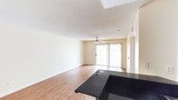 This single-story space offers an open and functional layout that blends the living and kitchen areas seamlessly. The living area is finished with warm hardwood-style flooring that extends throughout, contributing to a cohesive and inviting atmosphere. A ceiling fan is centrally mounted, providing both light and airflow to the space. Natural light enters generously through a sliding glass door fitted with vertical blinds, which also provides direct access to an outdoor area. The walls are painted in a neutral, light beige tone that enhances the brightness of the room.

The textured popcorn ceiling adds a classic architectural detail consistent throughout the space. A partial view of the kitchen reveals dark granite countertops that contrast elegantly with the lighter tones of the surrounding space, hinting at a well-appointed kitchen area. The transition between the kitchen and living space is defined by a countertop overhang, suggesting a convenient bar-style arrangement. Neutral baseboards frame the flooring cleanly throughout. Overall, the space presents a straightforward, functional design with practical finishes suited for everyday living.