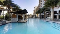 This impressive outdoor resort pool area combines elegant design with a welcoming, vacation-ready atmosphere. At its centerpiece is a generously sized swimming pool with crystal-clear turquoise water, designed to accommodate both leisurely lounging and active recreation. The pool's smooth, curving edges and shallow-entry sections add to its resort-caliber appeal.

Flanking the pool are private cabanas featuring sleek, modern canopies with flowing white curtains, stone accent walls, and comfortable seating — ideal for guests seeking shade and seclusion. Tall, swaying palm trees are thoughtfully interspersed throughout the deck, enhancing the tropical ambiance and providing natural greenery against the hardscape.

The surrounding pool deck is outfitted with lounge chairs and umbrellas, offering ample space for relaxation. In the background, a grand multi-story building rises with a warm, Mediterranean-inspired façade of arched windows, balconies, and earth-toned finishes, framing the outdoor space with architectural grandeur.

A pavilion-style structure at the far end of the pool anchors the space and appears to serve as an additional gathering or entertainment area. The overall setting evokes a high-end resort experience, blending sophisticated architecture, lush landscaping, and thoughtfully curated amenities into a cohesive and inviting outdoor environment.