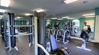 This fitness center offers a fully outfitted workout environment designed to accommodate a wide range of exercise routines. The space is equipped with multiple cardio machines, including elliptical trainers and treadmills, providing ample options for cardiovascular conditioning. A dedicated strength training station anchors one section of the floor, offering users a versatile platform for resistance-based workouts.

Full-length mirrors line the walls, visually expanding the space and allowing users to monitor their form throughout their sessions. The rubber flooring throughout provides both durability and comfort underfoot, making it suitable for a variety of training activities. Overhead fluorescent lighting ensures the space is evenly and brightly illuminated.

The layout is thoughtfully organized to allow clear movement between equipment, reducing congestion during peak hours. A textured ceiling completes the utilitarian aesthetic, keeping the focus firmly on function. Natural light filters in through windows visible in the mirrored reflections, adding a welcoming brightness to the environment. Overall, this fitness center is a practical and well-maintained space suited to meet the everyday workout needs of its users.