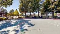 The West Side Tennis Courts offer a well-appointed outdoor recreational facility featuring multiple hard-surface tennis courts finished in a classic green and gray color scheme with clearly marked white court lines. The courts are fully enclosed by sturdy chain-link fencing, providing both safety and a defined play area, while gated entry points allow convenient access from multiple sides.

Tall, mature trees surround the perimeter of the facility, offering natural shade and a pleasant, park-like atmosphere. Manicured shrubs and landscaped planting beds line the fencing, adding greenery and visual appeal to the setting. The courts benefit from dedicated overhead lighting mounted on tall poles, ensuring usability during evening hours.

A solid teal-colored practice backboard is positioned along one end of the facility, ideal for solo practice sessions. Seating is available courtside, providing a comfortable spot for players to rest between games. The surrounding environment includes adjacent residential buildings and open green space, contributing to a welcoming neighborhood atmosphere. Under open skies, the facility is bathed in natural sunlight, making it an inviting destination for players of all skill levels throughout the day.
