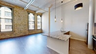 This thoughtfully converted urban loft showcases a seamless blend of industrial character and contemporary design across a single open floor plan. Original exposed brick walls anchor the living area, complemented by large arched windows that flood the space with natural light and frame views of the surrounding cityscape. Structural columns and exposed ductwork preserve the building's historic industrial roots, while a wood-paneled ceiling adds warmth and texture overhead.

The open-concept kitchen is a standout feature, equipped with stainless steel appliances including a refrigerator, electric range, and over-the-range microwave. Sleek dark cabinetry pairs elegantly with granite countertops, and a kitchen island with an integrated sink provides additional workspace and casual dining space. Stylish pendant lighting with brass accents hangs above the island, adding a refined touch.

The living area benefits from generous ceiling height and light hardwood-style flooring that extends throughout the space, creating a cohesive and airy atmosphere. Wall-mounted sconces along the hallway provide subtle accent lighting leading to the bathroom, which features a walk-in shower, accessibility grab bars, and consistent wood-look flooring. The overall ambiance is a sophisticated balance of raw, historic character and polished modern living.