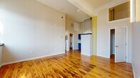 This single-floor loft-style unit showcases an open, airy layout defined by soaring ceilings with exposed white beams that lend an industrial character to the living space. Rich, warm-toned hardwood floors run throughout, adding both texture and visual continuity across every room. The expansive main living area benefits from generous wall space and excellent ceiling height, creating an open and breathable atmosphere.

The kitchen is a standout feature, equipped with bold blue-grey cabinetry extending to the upper walls, white countertops, a stainless steel dishwasher and refrigerator, a subway tile backsplash, and industrial-style pendant lighting. A partial partition wall with a peninsula counter separates the kitchen from the living area while maintaining an open feel.

The bedroom offers a quiet retreat with a large roller-shade window framing city skyline views, filling the room with natural light. A separate, smaller room provides flexible use as an office or additional sleeping space, also with a city-facing window.

The bathroom is finished with large-format white subway tile surrounding a full soaking tub, complemented by matte black fixtures for a clean, contemporary contrast. Throughout the unit, details such as the yellow accent column, track lighting hardware, and city views contribute to a distinct urban loft ambiance.
