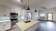 This single-story residence showcases a thoughtfully designed open-concept layout with a contemporary aesthetic throughout. The kitchen serves as the centerpiece, featuring gray shaker-style cabinetry extending to the ceiling, light quartz countertops, and a teal subway tile backsplash. A generously sized island with an undermount sink and matte black faucet anchors the space, illuminated by a pair of striking dark pendant lights. Stainless steel appliances, including a French door refrigerator, gas range, and built-in microwave, complete the culinary space. Wood-look flooring extends through the main living areas, while a ceiling fan adds comfort to the adjoining living space.

The bedrooms offer plush carpeting, tall ceilings, and natural light through windows including arched accent windows that add architectural character. The primary bathroom features dark countertops, white cabinetry, large-format tile flooring, a soaking tub, and a separate tiled shower. A spacious walk-in closet with wire shelving provides ample storage. An additional bathroom with similar finishes serves the secondary rooms, completing this well-appointed residence.