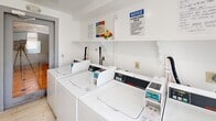 This shared laundry facility offers a clean, efficiently organized space designed for resident use. The room features multiple commercial-grade top-loading washing machines lined along one wall, complemented by stacked dryer units that maximize the use of vertical space. Each machine is coin-operated and clearly labeled, making the laundry process straightforward for users. The walls are painted white, contributing to a bright and tidy atmosphere, while light-toned tile flooring adds to the room's clean aesthetic. A wall-mounted ventilation fan near a window ensures adequate airflow and helps manage heat and humidity generated by the machines. Natural light filters in through the window, enhancing the overall brightness of the space. A shelf above the washers provides a convenient surface for detergent or personal items during laundry cycles. Posted notices and laundry room rules are clearly displayed on the walls, keeping residents informed of proper usage policies. The room is accessed through a standard door, and its compact yet functional layout ensures all essential laundry amenities are within easy reach, making it a practical and well-maintained communal resource.