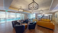 This thoughtfully designed single-floor amenity space offers a seamless blend of comfort and contemporary style across several distinct yet interconnected areas. The main lounge serves as the heart of the space, featuring rich hardwood flooring, bold geometric pendant chandeliers, and a curated mix of navy blue swivel chairs, vibrant mustard yellow sofas, and cream accent seating. A striking natural stone fireplace wall with a sleek linear hearth and built-in display shelving anchors one end of the lounge, while a large-format display screen adds a modern focal point. Adjacent to the lounge, a well-equipped kitchenette boasts dark tile backsplash, stainless steel appliances, white cabinetry, and a bar-height counter with seating. Glass partitions open to a bright indoor pool area, with abundant natural light filtering through large windows. Horizontal shiplap-style wall paneling and natural stone accent walls appear throughout, lending warmth and texture to the corridors and transitional spaces. The entry vestibule is clean and modern, with wood-toned wall cladding and digital display screens. Glass-railed stairways with wood handrails connect different levels of the entry zone, adding architectural interest. Throughout, recessed lighting and wall sconces create a welcoming and polished atmosphere.