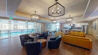 This thoughtfully designed single-floor amenity space offers a seamless blend of comfort and contemporary style across several distinct yet interconnected areas. The main lounge serves as the heart of the space, featuring rich hardwood flooring, bold geometric pendant chandeliers, and a curated mix of navy blue swivel chairs, vibrant mustard yellow sofas, and cream accent seating. A striking natural stone fireplace wall with a sleek linear hearth and built-in display shelving anchors one end of the lounge, while a large-format display screen adds a modern focal point. Adjacent to the lounge, a well-equipped kitchenette boasts dark tile backsplash, stainless steel appliances, white cabinetry, and a bar-height counter with seating. Glass partitions open to a bright indoor pool area, with abundant natural light filtering through large windows. Horizontal shiplap-style wall paneling and natural stone accent walls appear throughout, lending warmth and texture to the corridors and transitional spaces. The entry vestibule is clean and modern, with wood-toned wall cladding and digital display screens. Glass-railed stairways with wood handrails connect different levels of the entry zone, adding architectural interest. Throughout, recessed lighting and wall sconces create a welcoming and polished atmosphere.