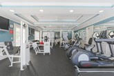 This fitness center in Phenix City, Alabama, offers a thoughtfully designed workout space that combines functionality with refined architectural character. The room is distinguished by elegant tray ceilings adorned with decorative crown molding and accented in a bold teal hue, lending the space a polished, upscale feel uncommon in typical gym environments. Durable rubber flooring throughout provides safety and comfort underfoot.

The space is comprehensively equipped for a full range of fitness activities. Cardio offerings include Star Trac stationary bikes, elliptical trainers, and treadmills arranged to maximize floor space. For strength training, the facility features a Cybex cable machine, a multi-station weight system, a free-weight dumbbell rack, a power rack with barbell and weight plates, and a selection of weight benches. Additional functional equipment such as stability balls rounds out the workout options.

Natural light filters in through multiple windows fitted with slatted blinds, creating a bright and energizing atmosphere. Full-length mirrors along one wall expand the sense of space and allow for form monitoring during exercise. Ceiling fans and recessed lighting ensure comfort and visibility throughout. A wall-mounted flat-screen television adds an entertainment element to workouts. The facility also includes clearly marked exit access, reflecting attention to safety standards.