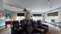 The Club Room is a well-appointed community gathering space designed to accommodate a variety of social activities. Rich, dark hardwood flooring flows throughout the entire space, anchoring the warm, neutral taupe and gray color palette. The kitchen area serves as a central hub, featuring dark cabinetry with ample storage, light quartz countertops, stainless steel appliances including a full-sized refrigerator and microwave, and a spacious island with seating for multiple bar-height chairs. Pendant lighting hangs above the island, adding focused illumination and a modern aesthetic touch. Recessed ceiling lights provide consistent brightness throughout.

Adjacent to the kitchen, a game and entertainment zone includes a full-sized billiards table and a wall-mounted flat-screen television, making it ideal for casual recreation. A dedicated lounge area offers generous natural light through large windows with views of the surrounding grounds. This seating zone features a plush sofa, coordinating blue accent chairs, a wood coffee table, and a patterned area rug, creating a comfortable and inviting retreat. A secondary seating arrangement with a wall-mounted TV and a dining table with blue chairs provides additional gathering options. The overall layout supports both lively group events and relaxed everyday use.