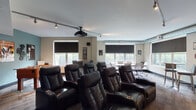 The Club Room is a well-appointed community gathering space designed to accommodate a variety of social activities. Rich, dark hardwood flooring flows throughout the entire space, anchoring the warm, neutral taupe and gray color palette. The kitchen area serves as a central hub, featuring dark cabinetry with ample storage, light quartz countertops, stainless steel appliances including a full-sized refrigerator and microwave, and a spacious island with seating for multiple bar-height chairs. Pendant lighting hangs above the island, adding focused illumination and a modern aesthetic touch. Recessed ceiling lights provide consistent brightness throughout.

Adjacent to the kitchen, a game and entertainment zone includes a full-sized billiards table and a wall-mounted flat-screen television, making it ideal for casual recreation. A dedicated lounge area offers generous natural light through large windows with views of the surrounding grounds. This seating zone features a plush sofa, coordinating blue accent chairs, a wood coffee table, and a patterned area rug, creating a comfortable and inviting retreat. A secondary seating arrangement with a wall-mounted TV and a dining table with blue chairs provides additional gathering options. The overall layout supports both lively group events and relaxed everyday use.