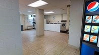 This shared laundry facility is a practical, well-organized space designed to accommodate multiple users at once. The open floor plan is anchored by structural columns and finished with light-toned ceramic tile flooring throughout, making for easy maintenance and a clean appearance. The drop ceiling is equipped with fluorescent panel lighting that ensures bright, even illumination across the entire area. Along the walls, a row of top-load washing machines and front-load dryers are arranged for efficient use, with stacked dryer units also present to maximize capacity. Informational signage is posted throughout, including laundry usage guidelines and alerts to assist users. A baseboard heating unit is visible along one wall, providing warmth during colder months. The structural columns divide the space subtly without impeding circulation, and the layout allows for comfortable movement between machines. Overall, the facility is straightforward and utilitarian in design, prioritizing function and accessibility as a communal laundry resource.