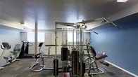 This fitness center offers a practical and well-equipped workout environment designed to support a range of strength training and exercise routines. The space is anchored by a large, multi-station cable machine constructed of heavy-gauge steel, featuring multiple weight stacks, pulleys, and attachment points to accommodate a wide variety of exercises. Surrounding the central cable system are additional strength training stations, including a dedicated bench and other specialized machines, providing users with a comprehensive workout experience. The flooring is finished in a durable, dark carpet suited for high-traffic fitness use. Walls are painted in a cool blue-gray tone, lending a focused and energetic atmosphere to the space. Natural light filters in through windows along the perimeter, complementing the overhead lighting to keep the environment bright and functional. A wall-mounted mirror reflects the equipment, enhancing the sense of openness within the room. Signage on the walls provides facility guidelines for users. The single-floor layout ensures easy accessibility, and the overall design prioritizes utility and efficiency, making it a well-rounded space for residents or members looking to maintain an active lifestyle.