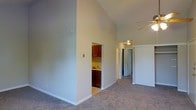 This single-story residence showcases a spacious, open layout accentuated by dramatically vaulted ceilings that lend an airy, expansive feel to the main living area. The living space is finished with plush wall-to-wall carpeting in a neutral tone, contributing to a comfortable and inviting atmosphere. Smooth, light gray walls complement the overall clean aesthetic throughout.

A ceiling fan is mounted overhead, providing both functional air circulation and a subtle design element. A partial wall with a cutout opening provides a glimpse into the kitchen, which features rich, dark wood cabinetry and a sink set against tile flooring — a practical contrast to the carpeted living space.

A hallway extends from the main living area toward additional rooms, with doors visible at the far end suggesting access to bedrooms and at least one bathroom. The corridor is well-lit by ceiling-mounted lighting, maintaining continuity with the bright, neutral palette found throughout the home. The overall design is straightforward and functional, with the vaulted ceilings serving as the most notable architectural feature of the space.