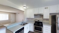 This single-floor space showcases a well-appointed kitchen that serves as a central highlight. The kitchen features crisp white cabinetry extending to the upper walls, providing generous storage throughout. Light-toned laminate countertops run along an L-shaped configuration, offering ample workspace for meal preparation. A double-basin stainless steel sink is positioned along the counter peninsula, complemented by a modern brushed-nickel faucet. Stainless steel appliances include a gas range with oven and a dishwasher, lending a contemporary, cohesive look to the space. An under-cabinet range hood is installed above the stove for ventilation. Dark hardwood-style flooring grounds the kitchen and contrasts neatly with the light cabinetry and counters. Flush-mount ceiling lighting provides even illumination throughout. A pass-through opening connects the kitchen to an adjacent room, enhancing the sense of openness and allowing for easy flow between spaces. The overall ambiance is clean, bright, and functional, with a neutral color palette that creates a versatile, move-in-ready environment.