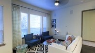 This single-floor space centers around a thoughtfully designed living area that balances comfort and contemporary style. Large windows fitted with horizontal blinds and patterned draping panels allow generous natural light to fill the room, creating a bright and welcoming atmosphere. The ceiling is finished in a deep blue-gray tone, adding an unexpected design element that contrasts with the crisp white walls and contributes to the room's visual depth.

The seating arrangement features a pair of navy blue armchairs positioned near the windows, complemented by a white upholstered sofa accented with houndstooth and geometric throw pillows in black and white. A circular glass-top coffee table anchors the central seating area, while a low-profile media console along the back wall provides functional storage. A bold black-and-white diamond-patterned area rug defines the living space and adds graphic energy underfoot.

Lighting includes a ceiling-mounted fixture paired with a sleek arc floor lamp positioned near the console, offering layered illumination. Framed artwork displayed in a vertical arrangement on one wall adds a curated, gallery-like touch, completing the space's polished yet livable character.