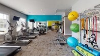 Located at 2640 Latrobe Ave in Fayetteville, NC, this single-floor fitness center is designed to support a comprehensive workout experience. The space is visually energized by bold teal and yellow-green accent walls that contrast with neutral gray tones, creating a motivating atmosphere throughout. Durable, patterned carpet tile flooring covers the workout areas, and recessed lighting combined with an abundance of natural light from multiple large, blinds-fitted windows keeps the space bright and welcoming.

The main cardio area is lined with a wall of floor-to-ceiling mirrors and equipped with a row of treadmills, elliptical trainers, and stationary bikes, each paired with mounted flat-screen TVs for entertainment during workouts. A dedicated strength training section houses a multi-function cable machine, adjustable weight benches, a plate-loaded rack, and a well-stocked dumbbell rack spanning a wide range of weights. Additional functional fitness equipment includes medicine balls, stability balls, and a captain's chair for core training.

The layout flows openly between cardio and strength zones, with a clear passage connecting the two areas. Windows throughout the space offer views of an outdoor pool area, adding a refreshing visual element. The overall design balances functionality and aesthetics, making this fitness center a thorough and inviting facility.