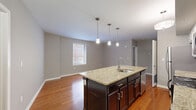 Located at 459 Locust Ave in Charlottesville, VA, this two-bedroom, two-bathroom residence offers a well-designed single-floor layout. Rich hardwood flooring flows throughout the main living areas and bedrooms, creating a warm and cohesive aesthetic. The open-concept kitchen and living area serves as the heart of the home, centered around a striking dark-cabineted island with granite countertops, an undermount sink, and pendant lighting overhead. The kitchen features white upper cabinetry, a white subway tile backsplash, and a full suite of stainless steel appliances, including a refrigerator, range, and microwave. The adjoining living area is bright and open, with natural light filtering in through windows that frame views of the surrounding brick neighborhood. Both bedrooms offer generous proportions with the same warm hardwood floors and neutral gray walls, and the primary bedroom provides direct access to a closet and bathroom. Each bathroom is finished with granite-topped vanities, dark cabinetry, and tile flooring, with one featuring a glass-enclosed walk-in shower. A dedicated laundry area with a stacked washer and dryer adds everyday convenience to this well-appointed Charlottesville residence.