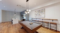 Located at 140 Rantoul Street in Beverly, Massachusetts, this thoughtfully designed community lounge offers a sophisticated blend of modern amenities and warm, inviting aesthetics across a single open floor. The space is unified by rich hardwood flooring throughout and a carefully curated mix of textures and finishes that create a cohesive, upscale atmosphere.

The game area serves as a central focal point, anchored by a handsome dark wood billiards table illuminated by a striking multi-globe pendant light fixture. A live-edge wood bar ledge with industrial-style stools runs along one wall, accompanied by a large abstract artwork, providing a casual perch for spectators. Adjacent to the game area, a full kitchenette features sleek gray cabinetry, stainless steel appliances including a built-in microwave, a white quartz waterfall island with leather bar seating, and a bold patterned accent wall that adds visual depth.

The lounge area is anchored by plush sectional and accent seating arranged around a large-screen television mounted on a shiplap feature wall. A distinctive slatted wood tray ceiling with a sculptural sputnik chandelier adds architectural character overhead. A shuffleboard table and large floor-to-ceiling windows flood the space with natural light and offer views to an outdoor patio area. A vintage Casa de Lucca restaurant sign serves as a charming nod to the building's local history.