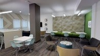 This single-floor communal lounge is thoughtfully designed to encourage both social interaction and focused work. The space features a versatile open layout with multiple distinct seating zones, allowing occupants to choose settings that best suit their needs. A standout green bench runs along one wall, paired with light teal molded chairs and a geometric-patterned acoustic wall panel that adds both visual interest and sound absorption. Pendant lighting hangs from the white ceiling, complementing the recessed fixtures and providing well-distributed illumination throughout. A bold structural column in dark charcoal serves as an architectural focal point, anchoring the room while defining separate areas within the open plan. Patterned carpet in earthy tones adds texture underfoot and contributes to the space's warm yet contemporary aesthetic. A wall-mounted display screen and a small café-style table with shell chairs offer additional functional areas. Curated circular artwork adorns the walls, adding a refined, gallery-like quality. Natural light filters in through slatted window blinds, softening the atmosphere. Overall, the space balances modern design with practical amenities, making it well-suited as a collaborative lounge or common area.