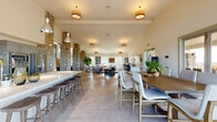 This single-floor space in Folsom, California, is defined by its striking architectural design and open, airy layout. Soaring vaulted ceilings amplify the sense of volume throughout, while light herringbone-patterned flooring flows seamlessly from zone to zone, creating visual continuity across the entire space.

Dramatic natural stone columns serve as the architectural centerpiece, rising from floor to ceiling and lending a warm, organic texture that contrasts beautifully with the clean white walls. Multiple lounge areas are thoughtfully arranged, furnished with comfortable sofas, upholstered armchairs, and area rugs, accented by large indoor plants that enliven the atmosphere.

A long white bar counter with bar stools defines the dining and gathering area, complemented by a substantial communal wood dining table nearby. Warm amber pendant lighting illuminates these zones, adding a welcoming glow.

The fully equipped kitchen features stainless steel appliances, gray cabinetry, open shelving, a white tile backsplash, and a large island with seating—ideal for event catering or group use. A dedicated workstation area is visible near the stone columns. Large windows invite natural light throughout, and glass doors provide access to outdoor areas, further enhancing the bright, open ambiance of this well-appointed space.