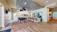 Located in Saint Johns, FL, this single-floor community clubhouse offers a generously sized, thoughtfully designed gathering space ideal for socializing, recreation, and relaxation. The main game and lounge area is the centerpiece of the space, boasting soaring vaulted ceilings clad in rustic wood planking, a ceiling fan, and large glass doors that flood the room with natural light and provide views of the outdoor amenities. A full-size pool table anchors the space atop a vibrant patterned area rug, while comfortable sectional seating, wall-mounted TVs, and decorative elements like Moroccan pendant lights and coastal artwork create an inviting atmosphere. A stylish bar-height island with a quartz countertop and colorful accent chairs connects to a coffee and beverage station framed by teal subway tile, teal cabinetry, and pendant lighting. The wide, art-lined corridor leads to additional seating areas, including a cozy reading nook with booth-style seating and a bookshelf. Textured wallcoverings, bold patterned carpet, wood-look flooring, and curated artwork throughout give the space a polished, resort-inspired aesthetic. A dedicated lounge area with a large sectional sofa, oversized chandelier, and board games completes this well-appointed clubhouse.