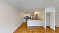 This single-floor space is thoughtfully designed with a clean, modern aesthetic throughout. The open-concept layout connects the kitchen and living areas seamlessly, anchored by warm-toned hardwood flooring that runs throughout the main living spaces. The kitchen is well-equipped with stainless steel appliances, including a french-door refrigerator, electric range with microwave, and features crisp white cabinetry paired with a mosaic tile backsplash and neutral countertops. A peninsula with pendant lighting in a brushed gold finish provides additional prep space and casual seating. The living area is generously proportioned and bathed in natural light from large windows and a sliding glass door that opens to the outdoors. A built-in closet offers convenient storage within the living space. The bathroom is finished with tile flooring, a tub-shower combination with tile surround, a dark wood vanity with a white countertop, and a stacked washer and dryer unit, consolidating laundry within the bathroom for added convenience. Recessed lighting and bright white walls contribute to the overall light and airy ambiance of the space.
