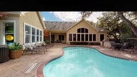 This single-story community clubhouse and outdoor amenity space is designed to offer a well-rounded recreational experience. The building itself features a classic stucco exterior with a pitched shingle roof and a striking arched window facade that allows natural light to pour into the interior. Large multi-pane windows line the structure, maintaining a visual connection between the indoor and outdoor spaces.

The centerpiece of the outdoor area is a generously sized freeform swimming pool with a brick-trimmed surround and clear turquoise water. The pool is complemented by an expansive stamped concrete patio that wraps around both sides of the building, providing ample space for lounging and socializing. Multiple wrought iron chaise lounges are arranged throughout the deck, ideal for relaxing poolside.

A dedicated outdoor dining area features a cast iron patio table with matching chairs, perfectly suited for outdoor gatherings. An outdoor grill station adds convenience for al fresco dining. The entire outdoor space is enclosed by a wrought iron fence, offering both security and open views of the surrounding mature oak trees and lush landscaping. The natural canopy of trees creates a shaded, serene atmosphere that enhances the overall ambiance of this inviting outdoor retreat.
