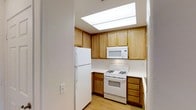 This single-floor space centers around a compact, efficiently designed kitchen that makes excellent use of its footprint. A standout architectural feature is the overhead skylight, which floods the kitchen with abundant natural light, creating a bright and airy atmosphere throughout the day.

The kitchen is lined with warm-toned, medium oak wood cabinetry that extends to the upper and lower sections of the walls, providing generous storage space. The upper cabinets are arranged symmetrically, framing the skylight and maximizing vertical storage capacity. Light-colored countertops contrast cleanly against the wood tones, contributing to a balanced, cohesive aesthetic.

White appliances, including a gas range, over-the-range microwave, and a full-sized refrigerator, are neatly integrated into the layout, offering a full suite of cooking and food storage functionality. The gas range is centrally positioned along the back wall, flanked by the microwave above and ample counter space on either side.

Light hardwood-style flooring runs through the space, complementing the warm wood cabinetry and adding continuity throughout. The overall ambiance is clean, practical, and well-illuminated.
