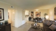 This single-floor unit showcases an open-concept layout that seamlessly connects the living, dining, and kitchen areas into one cohesive space. Light hardwood-style flooring runs throughout, lending warmth and a sense of continuity across the entire floor plan.

The living area is anchored by a round wooden coffee table set atop a textured area rug, creating a comfortable and inviting atmosphere. Crown molding lines the ceiling, adding a refined architectural detail to the otherwise clean, neutral interior.

The dining area accommodates a full rectangular table with seating for several guests and is accented by a warm-toned abstract artwork mounted on the wall, contributing a decorative focal point to the space.

The kitchen is partially visible from the main living area and features white cabinetry paired with a mosaic tile backsplash, offering a subtle contrast of texture. The bright, white finish of the kitchen reflects light effectively, keeping the space feeling open and airy.

A hallway near the entry leads to additional rooms, while multiple doors suggest access to private areas such as bedrooms or storage. The overall ambiance is bright, clean, and functional, with a neutral palette that offers versatility for personalization.