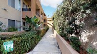 This multi-story residential complex showcases a classic Southern California architectural style, characterized by warm tan stucco exteriors, tile roofing accents, and black wrought-iron balcony railings. The buildings are arranged around a generous central courtyard, which serves as the communal heart of the property. Mature shade trees anchor the open-air patio, casting dappled light across the wide concrete walkways and creating a relaxed, park-like atmosphere.

Landscaping is a prominent feature throughout the property. Lush plantings—including tropical banana-leaf plants, ferns, ornamental shrubs, and towering shade trees—line the perimeter walkways and raised planting beds, offering a verdant, well-maintained environment. Concrete pathways connect the various wings of the complex, with stairwells providing access between levels.

Resident-facing amenities visible on the grounds include a vending machine station and clearly marked pet waste disposal areas, indicating a pet-friendly community. Individual units feature private balconies or patios, offering outdoor space and views of the landscaped surroundings. The overall ambiance is calm and well-tended, with abundant greenery softening the built environment and contributing to a comfortable, community-focused residential setting.