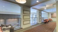 This single-floor space presents a well-appointed interior corridor or hallway area designed with both functionality and aesthetics in mind. The layout is open and airy, benefiting from recessed ceiling lighting complemented by a prominent overhead pendant fixture that illuminates the space evenly. Structural columns punctuate the corridor, adding architectural definition while framing the flow of movement through the area.

A notable design feature is the series of ornately framed rectangular panels mounted along the neutral-toned walls, providing visual interest and a polished, curated atmosphere. The walls are finished in a clean, light color palette that enhances the brightness of the space.

Glass and mesh safety railings line portions of the corridor, offering views into adjacent areas and contributing to a sense of openness. Windows along the perimeter allow natural light to filter in, with views of an exterior brick facade visible beyond. The flooring consists of patterned carpeting in warm earth tones, adding comfort and texture underfoot.

A seating area with upholstered chairs is partially visible at one end, suggesting the space may serve a transitional or communal function within a larger building.