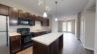 This single-floor residence offers a thoughtfully designed layout with two bedrooms, two bathrooms, and two closets connected by hallways. The open-concept kitchen and living area serves as the heart of the home, featuring rich dark-wood cabinetry that extends to the ceiling for ample storage, light countertops, and a central island with pendant lighting overhead. Black appliances — including a range, microwave, dishwasher, and stainless steel refrigerator — complement the kitchen's bold aesthetic. Track lighting and hardwood-style flooring flow throughout the main living area, which opens to a set of French doors that invite natural light and outdoor access. The living room is equipped with a ceiling fan for added comfort. Both bedrooms are carpeted and feature ceiling fans, with the primary bedroom offering generous proportions. The second bedroom also benefits from a large window with natural light. The bathrooms are finished with classic white subway tile surrounding a full soaking tub and shower combination, accented by chrome fixtures. A wide hallway lined with dark cabinetry connects the main areas to the private bedrooms, maintaining a clean and cohesive design throughout the space.