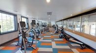 This fitness center is a single-level space designed to accommodate a comprehensive cardio-focused workout routine. The room is elongated, providing ample space for a generous lineup of equipment, including elliptical trainers, stationary bikes, treadmills, and rowing machines, all arranged in orderly rows along the length of the floor.

One of the most visually striking features of the space is the bold, geometric carpet tiles in a vibrant palette of orange, blue, gray, and white, adding a dynamic and energetic character to the room. The white ceiling is clean and modern, fitted with recessed lighting and ventilation units that ensure a comfortable environment during workouts.

Natural light filters in through windows along the left wall, while large glass panels on the right side offer views into an adjacent area, enhancing the sense of openness. Several of the cardio machines are equipped with personal monitors, adding a functional touch for users tracking their workouts. Overall, the space strikes a balance between practicality and contemporary design.