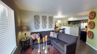 This single-floor residence offers a welcoming and well-appointed open-concept layout that connects the living, dining, and kitchen areas into one cohesive, functional space. The living area serves as the centerpiece of the home, anchored by a plush charcoal sectional sofa accented with decorative throw pillows in warm, complementary tones. A stylish coffee table with elegant candle holders adds a refined touch, while an abstract wall art trio lends the space an artistic character. Neutral gray walls and light ceilings create a clean, airy atmosphere throughout. The adjoining dining area features a dark wood table and chairs beneath a modern pendant light fixture, providing a defined yet open space for meals. The kitchen is visible beyond, featuring dark cabinetry, stainless steel appliances, and light countertops that strike a balance between style and practicality. A bright window and accent greenery bring natural elements indoors, contributing to the overall warmth of the space. The open floor plan ensures excellent flow between all areas, making the residence feel spacious and inviting. The cohesive design and careful attention to detail throughout create a comfortable and visually engaging living environment.