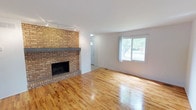 This two-story home offers a well-organized layout across both floors, combining classic design elements with practical living spaces. The main level is anchored by a generously sized living room, where warm-toned hardwood floors gleam beneath track lighting and a large front-facing window floods the space with natural light and outdoor views. A full-height brick fireplace with a substantial slate hearth serves as the room's defining architectural focal point, adding character and warmth to the open atmosphere.

Adjacent to the living room, the layout flows seamlessly toward the kitchen, which features light wood cabinetry, tile flooring, and a window that brings in additional natural light. The transition between the two areas is open and inviting, supporting a connected feel throughout the main level. A staircase leads to the upper floor, where additional rooms and a bathroom are arranged to provide private, comfortable spaces. The upper-level bathroom is equipped with standard fixtures and appears well-proportioned within the overall floor plan.

Throughout the home, the interplay of hardwood floors, white walls, and brick detailing creates a cohesive and timeless aesthetic that balances warmth with clean, understated design.