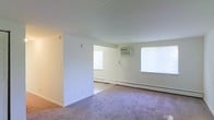 This single-floor residential space offers a clean, functional layout designed for comfortable everyday living. The interior features soft carpeted flooring throughout the main living area, providing a warm and comfortable underfoot feel. Neutral white walls and standard-height ceilings create a bright, open ambiance that maximizes the sense of space.

The living area flows into an adjoining room through an open half-wall partition, offering a sense of connected yet defined spaces. Natural light enters generously through multiple windows, keeping the interior airy and well-lit throughout the day. A wall-mounted air conditioning unit provides cooling comfort during warmer months, while baseboard heating units along the perimeter walls ensure efficient warmth during colder seasons.

The overall layout is straightforward and practical, with smooth transitions between areas. The light-toned walls and neutral flooring provide a versatile, move-in-ready backdrop suitable for a variety of interior styles and furnishing choices. The combination of natural light, climate control features, and open connectivity between rooms makes this a well-rounded and functional living space.