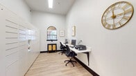 This single-floor business center is designed to provide a functional and stylish workspace for its users. The room features soaring ceilings that lend an open, airy feel, complemented by crisp white walls and light-toned wood-style flooring with dark baseboards that add a polished contrast throughout.

A long, wall-mounted white desk runs along one side of the room, accommodating multiple computer workstations complete with monitors, keyboards, and a printer, making it well-suited for productivity tasks. Ergonomic black office chairs are positioned at each station for comfortable use. An arched window centered on the far wall allows natural light to filter into the space, framing a decorative black cabinet with rattan-accented doors beneath it. Framed botanical artwork and a gold sunburst wall clock add tasteful decorative touches.

Along the opposite wall, a large bank of white package lockers provides secure parcel storage, a practical amenity for residents or building occupants. A glass-paneled dark-framed door connects directly to an adjacent fitness facility, offering convenient access to exercise equipment visible through the door. A skylight overhead further brightens the workspace, ensuring well-lit conditions throughout the day. Overall, the space strikes a balance between modern aesthetic design and practical, everyday functionality.