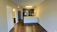 This single-floor unit showcases an open-concept layout that seamlessly connects the kitchen and living area, creating a spacious and functional environment. The kitchen is a standout feature, equipped with dark espresso cabinetry, granite countertops, and black stainless steel appliances, including a refrigerator, microwave, and range. A peninsula-style island provides additional counter space and serves as a natural dividing point between the kitchen and the main living area, with two pendant lights hanging above to enhance both task lighting and ambiance. A flush-mount ceiling fixture further illuminates the kitchen workspace. Rich, dark hardwood flooring flows continuously throughout the space, adding warmth and visual continuity. The neutral beige walls offer a versatile backdrop suitable for a variety of décor styles. A baseboard heating unit is positioned along the far wall, ensuring efficient climate control. Visible from the main living area, a hallway leads to additional spaces, including what appears to be a bathroom with a vanity and mirror, as well as an entry door. The overall design balances practicality with modern aesthetic appeal.