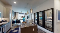 This thoughtfully designed single-floor residence offers a seamless blend of modern aesthetics and functional living. The open-concept layout connects the kitchen and living area, creating a spacious, inviting atmosphere. The kitchen is a standout feature, boasting a large island with a dark stone countertop, undermount sink, and elegant pendant lighting. Flat-panel cabinetry in a warm wood tone lines the walls alongside a full suite of stainless steel appliances, including a side-by-side refrigerator, electric range, microwave, and dishwasher. A built-in shelving nook near the entry provides convenient storage. The living area features a bold accent wall, wood-look flooring, and generous natural light from large windows dressed with sheer curtains. A glass-panel partition elegantly separates zones while maintaining visual openness. The bedroom is a serene retreat with plush carpeting, ample natural light, and high ceilings, complemented by a spacious walk-in closet with built-in shelving and hanging rods. A private en-suite bathroom features a granite-topped vanity, framed mirror, and a full tub-shower combination with wood-toned tile surround. A second bathroom with similar finishes provides added convenience.