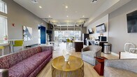 Located at 9050 W Chester Blvd in West Chester, Ohio, this single-floor community clubhouse offers a generously sized, thoughtfully designed gathering space that blends modern aesthetics with functional versatility. The interior is anchored by a soaring reception lounge featuring dramatic pendant lighting, light wood-look flooring, bold abstract artwork, and deep blue velvet accent chairs arranged around a marble-top table — creating a welcoming first impression.

The open-concept social lounge transitions seamlessly into a café-style amenity area equipped with a full-length bar counter, refrigerator, coffee station, and multiple wall-mounted televisions. A striking industrial-style overhead garage door opens fully to a covered outdoor terrace furnished with plush blue seating, a ceiling fan, and views of the surrounding community — effortlessly blending indoor and outdoor living.

A dedicated lounge zone features vibrant yellow-green bar stools, high-top tables, and cozy booth-style seating beside tall windows that flood the space with natural light. A private conference room, carpeted and outfitted with a long wooden table and plum-toned executive chairs, provides a quiet setting for meetings. Throughout, warm wood accent walls, recessed lighting, and a curated color palette of gray, magenta, and gold create a cohesive, upscale ambiance.